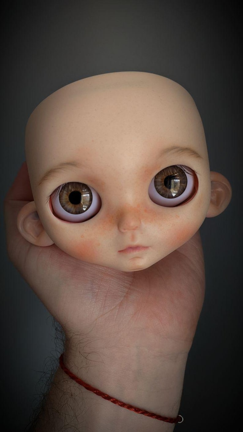 May include: A bald doll head with a smooth, pale skin tone, featuring large, round, brown eyes with detailed irises, subtle pinkish blushing on the cheeks, a small nose, and a neutral, closed-mouth expression with defined ears on the sides.