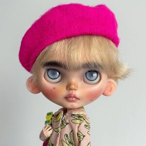May include: A doll with blonde hair and thick eyebrows wearing a bright pink beret. The doll has large, pale blue eyes, rosy cheeks, and a light complexion. It is dressed in a light pink collared jacket featuring a repeating yellow banana print.