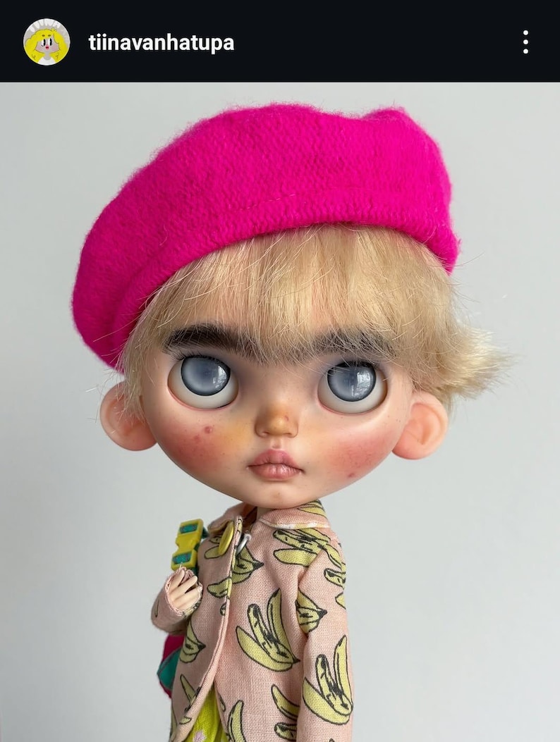 May include: A doll with blonde hair and thick eyebrows wearing a bright pink beret. The doll has large, pale blue eyes, rosy cheeks, and a light complexion. It is dressed in a light pink collared jacket featuring a repeating yellow banana print.