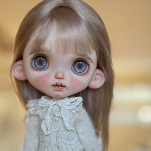 May include: A doll with long, straight, light brown hair and large, round, grey eyes with a glittery effect. The doll has a pale complexion with pink-tinted cheeks, wearing a cream-colored, long-sleeved, textured knit top with a lace collar.