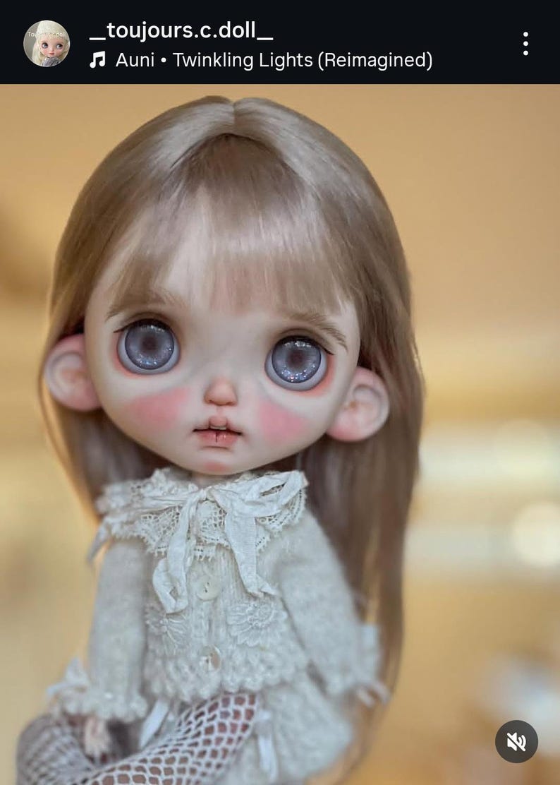May include: A doll with long, straight, light brown hair and large, round, grey eyes with a glittery effect. The doll has a pale complexion with pink-tinted cheeks, wearing a cream-colored, long-sleeved, textured knit top with a lace collar.