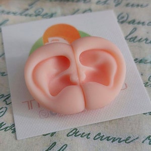 May include: A light peach-colored silicone mold featuring two symmetrical, detailed human ear impressions placed side-by-side. The mold is positioned on a small white square card with a partial orange circular logo visible underneath the object.