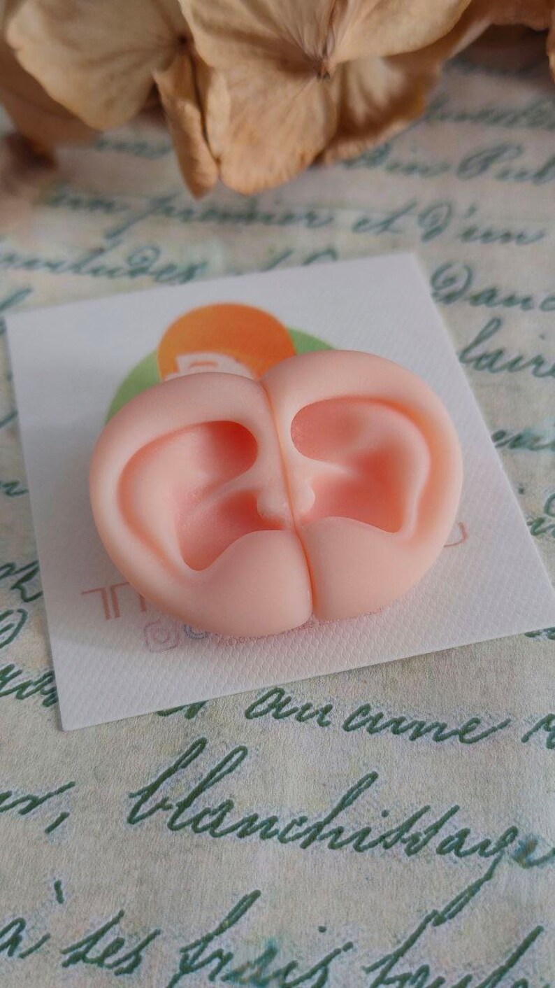 May include: A light peach-colored silicone mold featuring two symmetrical, detailed human ear impressions placed side-by-side. The mold is positioned on a small white square card with a partial orange circular logo visible underneath the object.