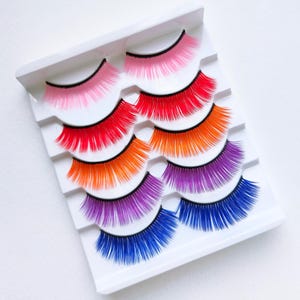 Könnte beinhalten: Ein Set bunter künstlicher Wimpern in einem weißen Plastikbehälter. Die Wimpern sind in Reihen angeordnet, wobei jede Reihe eine andere Farbe hat: rosa, rot, orange, lila und blau.