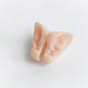 Può includere: Un paio di orecchie da elfo rosa pallido, progettate per essere indossate. Le orecchie presentano dettagli realistici dell'orecchio interno e sono realizzate in un materiale morbido e flessibile. Le orecchie sono su uno sfondo bianco.
