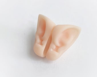 May include: A pair of pale pink elf ears, designed to be worn. The ears are detailed with realistic inner ear features and are made of a soft, flexible material. The ears are positioned on a white background.