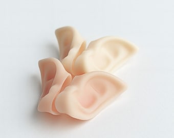 May include: Several prosthetic ear pieces in varying shades of pink and beige are arranged on a white surface. The ears are detailed with realistic folds and curves, suggesting a human ear shape. The lighting is soft, highlighting the textures.