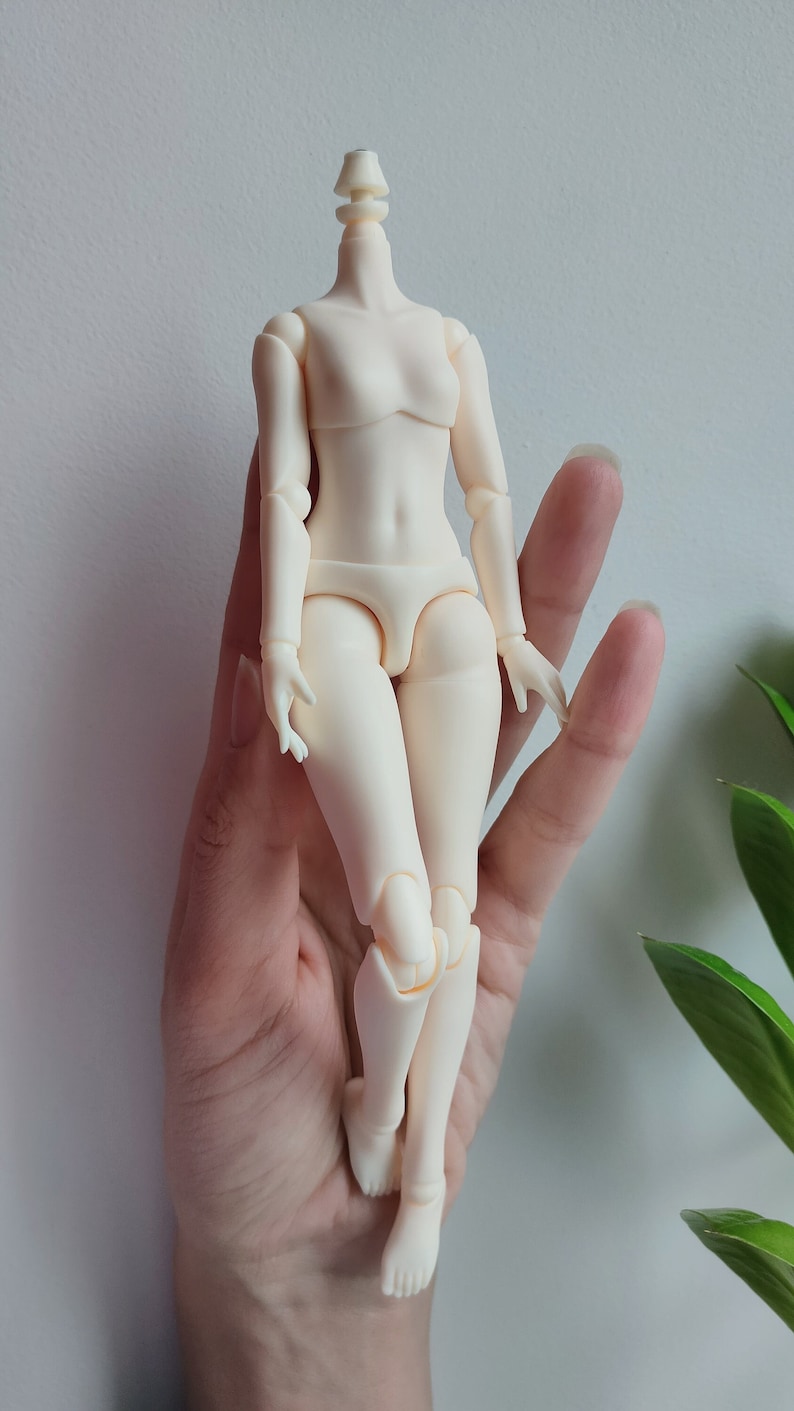 May include: A white plastic articulated doll body with no head. The doll is posed standing with its legs slightly bent. The doll is being held in a person's hand.