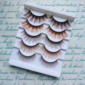 Könnte beinhalten: Ein Set mit sechs falschen Wimpern in einem weißen Plastiketui. Die Wimpern sind in drei Reihen mit jeweils zwei Wimpern angeordnet. Die Wimpern sind braun und haben ein natürliches Aussehen.