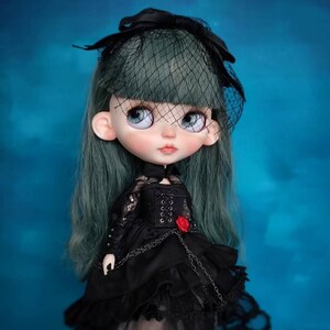 May include: A doll with long, muted green hair and a black veil headpiece. The doll wears a black dress featuring lace sleeves, a corset-style bodice, a small red rose accent at the waist, and a layered skirt with a delicate black chain detail.