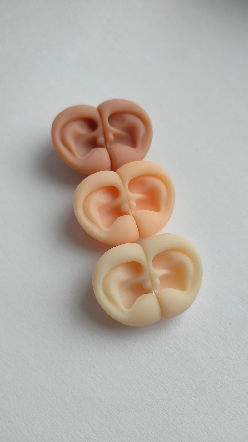 May include: Three pairs of small, molded ear-shaped objects arranged in a vertical column. Each pair is joined at the center, featuring realistic anatomical contours. The set displays a gradient of skin tones, ranging from a tan shade to a light beige.