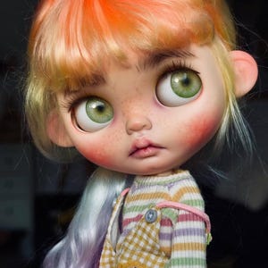 May include: A doll with large green eyes, freckles, and hair transitioning from orange to light purple. The doll wears a striped long-sleeve shirt under yellow and white checkered overalls featuring a pink graphic with the text RORD JUNK.