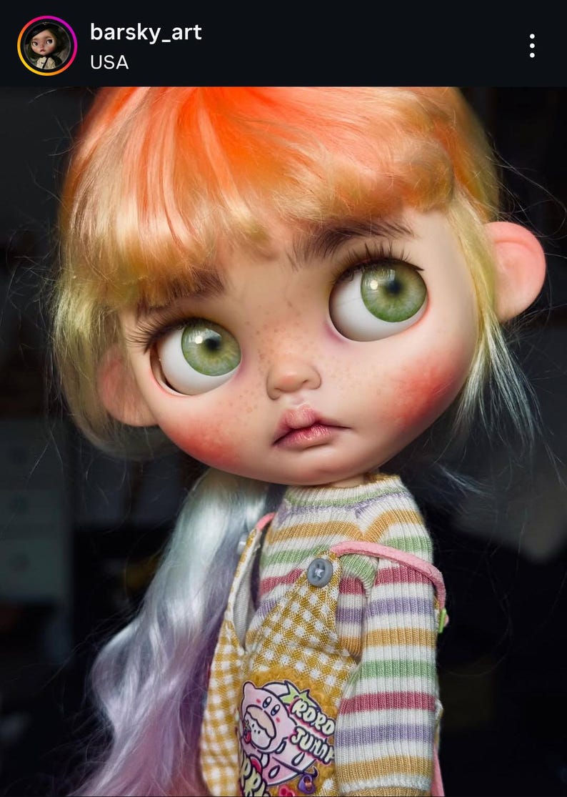 May include: A doll with large green eyes, freckles, and hair transitioning from orange to light purple. The doll wears a striped long-sleeve shirt under yellow and white checkered overalls featuring a pink graphic with the text RORD JUNK.