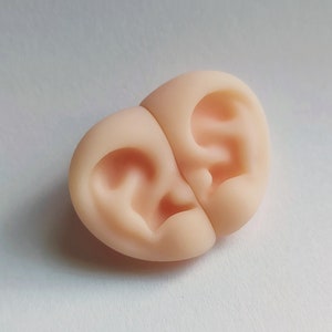 Könnte beinhalten: Ein Paar realistisch aussehende, hautfarbene Silikon-Ohrmuscheln. Die Formen sind so konzipiert, dass sie sich perfekt an das Ohr anschmiegen und zur Herstellung von individuellen Ohrstöpseln oder Hörgeräten verwendet werden.