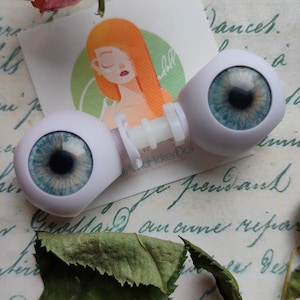 Könnte beinhalten: Ein Paar realistischer Puppenaugen mit blauen Iris und schwarzen Pupillen, verbunden durch einen weißen Kunststoffstab. Die Augen liegen vor einer quadratischen Karte mit einer Illustration einer Person mit orangefarbenem Haar und dem Text HitchHikerDoll.