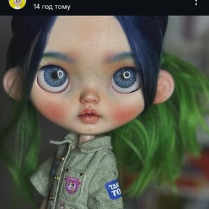 May include: A doll with large blue eyes, freckles, and two-toned hair featuring dark blue bangs and bright green sides. The doll wears a textured olive green short-sleeved shirt with small metallic buttons and several small patches, including one with the text TAK TO.