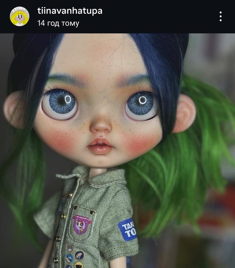 May include: A doll with large blue eyes, freckles, and two-toned hair featuring dark blue bangs and bright green sides. The doll wears a textured olive green short-sleeved shirt with small metallic buttons and several small patches, including one with the text TAK TO.