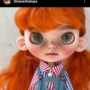 May include: A doll with bright orange hair, large green eyes, and freckles on the cheeks. The doll wears a red and white striped collared shirt under blue denim overalls featuring a heart-shaped cutout on the chest and a single white button.