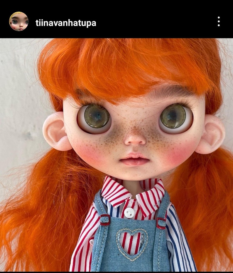 May include: A doll with bright orange hair, large green eyes, and freckles on the cheeks. The doll wears a red and white striped collared shirt under blue denim overalls featuring a heart-shaped cutout on the chest and a single white button.