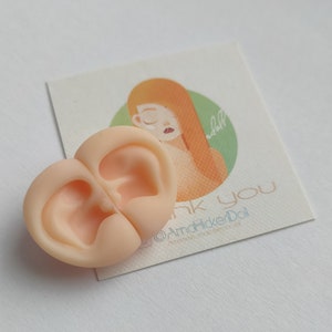 May include: A pair of light peach-colored silicone doll ears joined together. Behind the ears is a square card featuring an illustration of a person with long orange hair inside a green circle, with the text "thank you" and "@AmaFlickerDoll".
