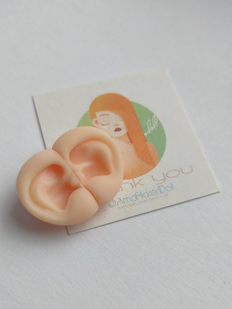 May include: A pair of light peach-colored silicone doll ears joined together. Behind the ears is a square card featuring an illustration of a person with long orange hair inside a green circle, with the text "thank you" and "@AmaFlickerDoll".