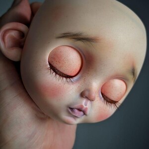 May include: A doll head with a smooth, pale complexion, featuring closed eyes with long dark eyelashes, subtle freckles across the nose and cheeks, soft pink blush, and delicate, sparse eyebrows. The head has a rounded shape and small ears.