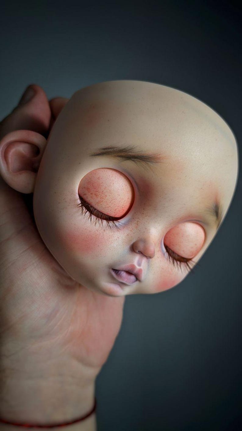 May include: A doll head with a smooth, pale complexion, featuring closed eyes with long dark eyelashes, subtle freckles across the nose and cheeks, soft pink blush, and delicate, sparse eyebrows. The head has a rounded shape and small ears.