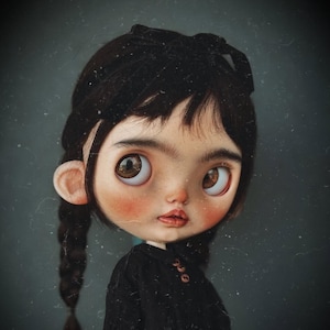 May include: A doll with a large head, brown eyes, and dark hair styled in two braids with a black bow. The doll has a pale complexion with rosy cheeks and wears a black long-sleeved shirt with small brown buttons at the collar area.