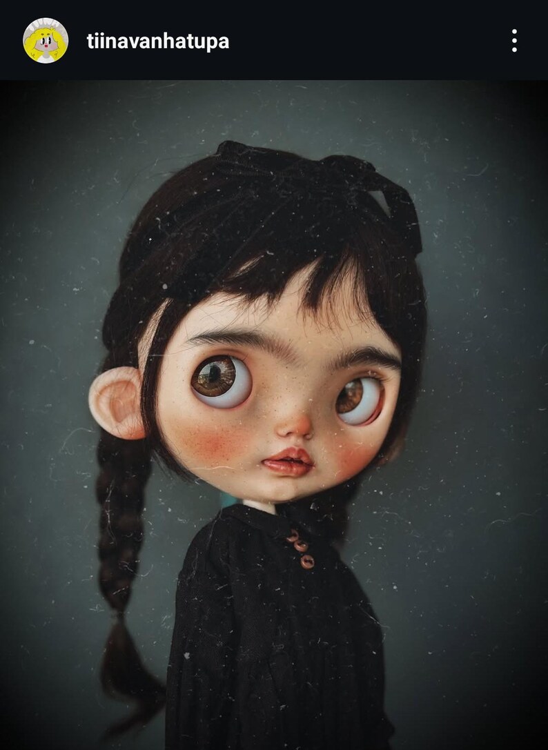 May include: A doll with a large head, brown eyes, and dark hair styled in two braids with a black bow. The doll has a pale complexion with rosy cheeks and wears a black long-sleeved shirt with small brown buttons at the collar area.