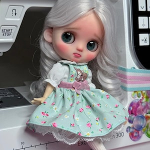 May include: A doll with long wavy silver hair, large teal eyes, and rosy cheeks. The doll wears a light green pinafore dress with a small deer graphic, pink floral patterns, and white lace trim, paired with white tights and light blue shoes.