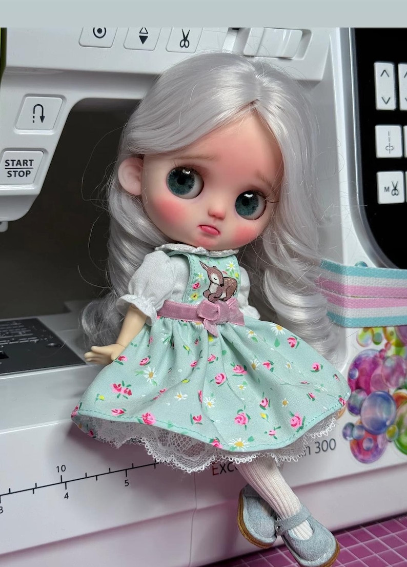 May include: A doll with long wavy silver hair, large teal eyes, and rosy cheeks. The doll wears a light green pinafore dress with a small deer graphic, pink floral patterns, and white lace trim, paired with white tights and light blue shoes.