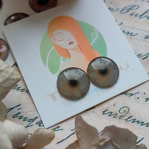 May include: Two grey and brown glass eyes with black pupils, on a white card with a green and orange illustration of a woman's face. The card has the text "The Doll" on it.