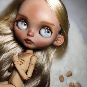 May include: A ball-jointed doll with long, straight blonde hair, large pale blue eyes, and a tan complexion. The doll has articulated limbs and is positioned with hands near the chest. Five additional interchangeable hand pieces are included.