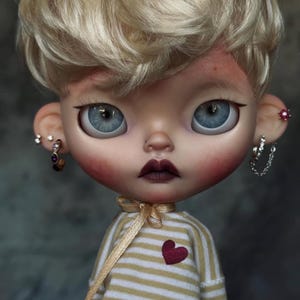 May include: A doll with short blonde hair, large blue eyes, and dark lips. The doll wears a beige and white striped long-sleeve shirt with a small red heart patch, a gold ribbon tie at the neck, and multiple silver-toned earrings in both ears.