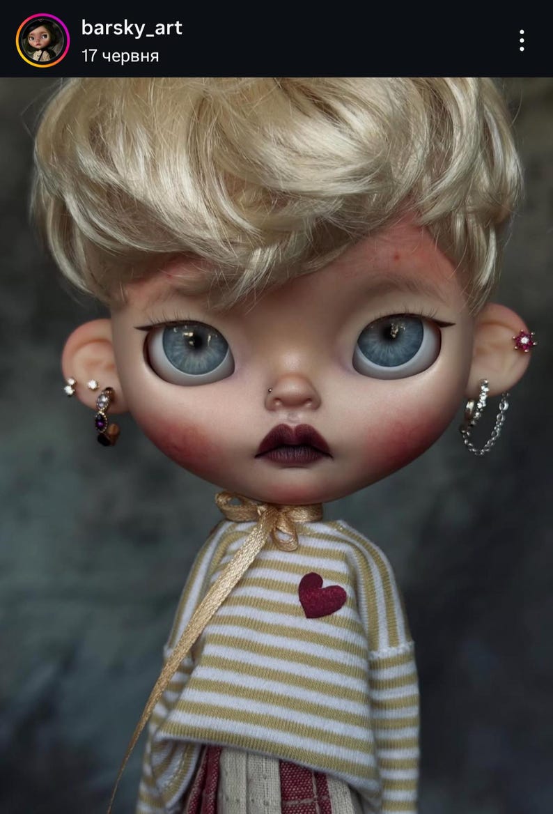 May include: A doll with short blonde hair, large blue eyes, and dark lips. The doll wears a beige and white striped long-sleeve shirt with a small red heart patch, a gold ribbon tie at the neck, and multiple silver-toned earrings in both ears.
