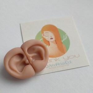May include: A pair of light peach-colored silicone doll ears positioned next to a square card featuring an illustration of a person with orange hair and the text "thank you @AmaHickerDoll handmade items for doll" printed in a light blue font.