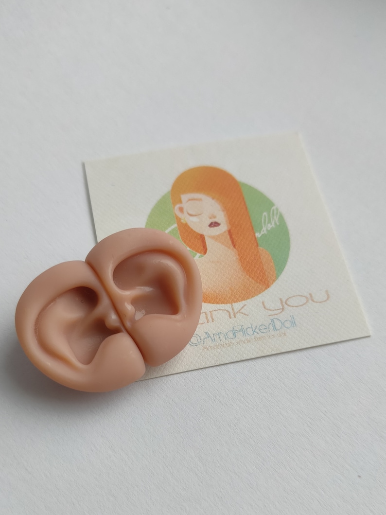 May include: A pair of light peach-colored silicone doll ears positioned next to a square card featuring an illustration of a person with orange hair and the text "thank you @AmaHickerDoll handmade items for doll" printed in a light blue font.