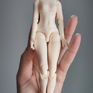 May include: A nude-colored articulated doll body with no head. The doll is standing on a hand.