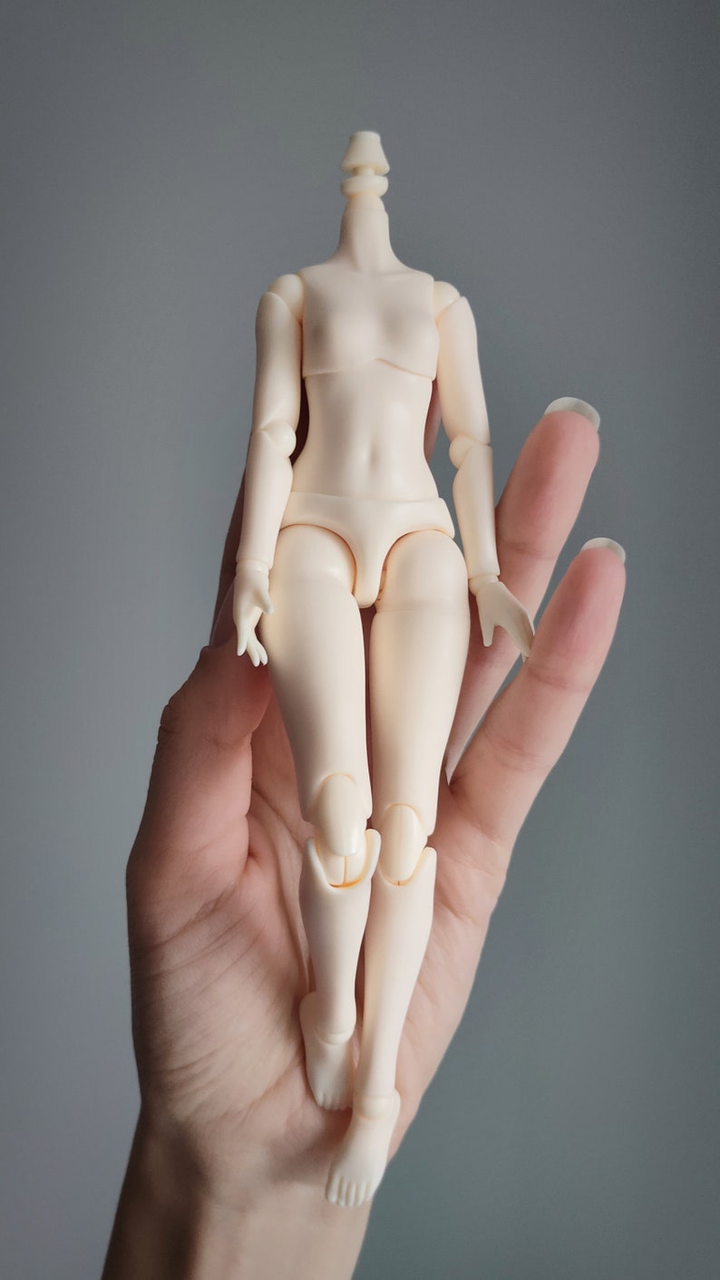 May include: A nude-colored articulated doll body with no head. The doll is standing on a hand.