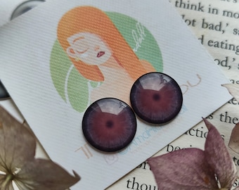 May include: Two purple and black doll eyes with a white iris and a black pupil. The eyes are on a white card with a cartoon illustration of a woman's face.
