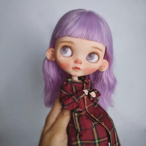 May include: A doll with shoulder-length lavender hair, large pale eyes, and small gold heart-shaped earrings. The doll is wearing a long-sleeved, button-down coat featuring a dark red, yellow, and black plaid pattern with a collar and cuffs.