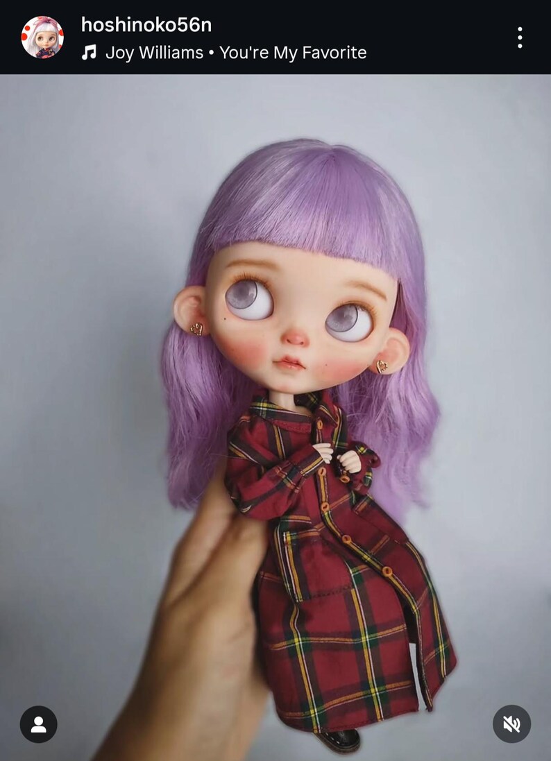 May include: A doll with shoulder-length lavender hair, large pale eyes, and small gold heart-shaped earrings. The doll is wearing a long-sleeved, button-down coat featuring a dark red, yellow, and black plaid pattern with a collar and cuffs.