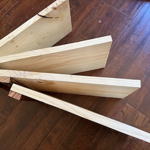 Op de afbeelding: Verschillende onafgewerkte houten planken van verschillende lengtes en breedtes, gerangschikt in een waaierpatroon. Het hout heeft een lichte, natuurlijke kleur met zichtbare nerven en knopen. De uiteinden van sommige planken hebben houten stukken bevestigd.