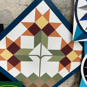 Puede incluir: Un bloque de colcha en forma de diamante con un borde azul. El centro del bloque presenta un tallo verde con dos conjuntos de patrones en forma de estrella naranja, amarillo y marr&oacute;n. La parte inferior del bloque tiene un patr&oacute;n de tri&aacute;ngulos marrones, verdes y grises.