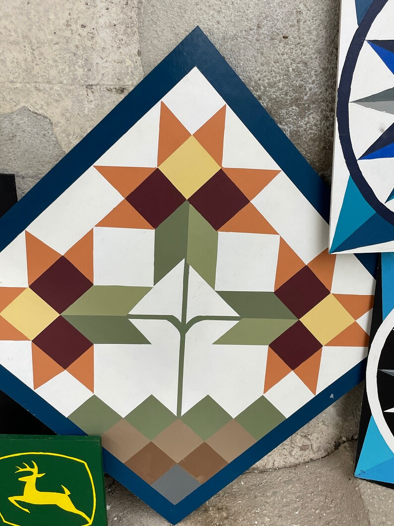 Puede incluir: Un bloque de colcha en forma de diamante con un borde azul. El centro del bloque presenta un tallo verde con dos conjuntos de patrones en forma de estrella naranja, amarillo y marr&oacute;n. La parte inferior del bloque tiene un patr&oacute;n de tri&aacute;ngulos marrones, verdes y grises.