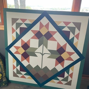 Puede incluir: Una pintura de granero cuadrada con un dise&ntilde;o geom&eacute;trico que presenta un patr&oacute;n de estrella en tonos de marr&oacute;n, rojo, amarillo y verde. La colcha est&aacute; enmarcada con un borde azul oscuro y un borde exterior verde.