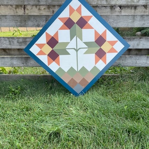 Puede incluir: Un letrero de madera en forma de diamante con un dise&ntilde;o de colcha que presenta tres girasoles estilizados en tonos naranja, amarillo, marr&oacute;n y verde. El fondo es de color azul oscuro.