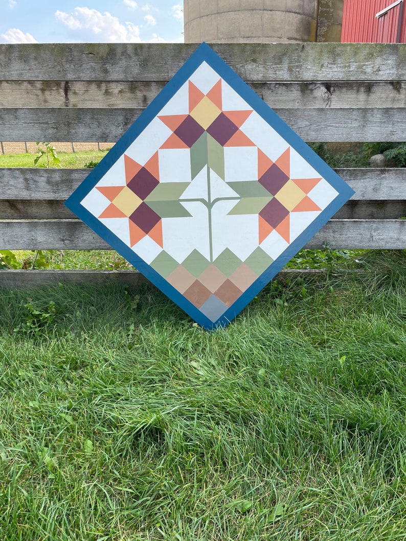 Puede incluir: Un letrero de madera en forma de diamante con un dise&ntilde;o de colcha que presenta tres girasoles estilizados en tonos naranja, amarillo, marr&oacute;n y verde. El fondo es de color azul oscuro.