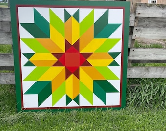 Puede incluir: Un bloque de colcha colorido con un diseño de estrella. El bloque está pintado en una tabla de madera y presenta tonos de verde, amarillo, naranja y rojo.
