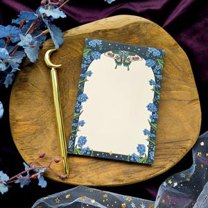 Puede incluir: Un bolígrafo dorado con una parte superior en forma de media luna descansa junto a un bloc de notas. El bloc de notas presenta un borde floral de flores azules y una ilustración de mariposa. Los artículos están sobre una bandeja de madera, con flores azules y una tela transparente con estrellas doradas en el fondo.